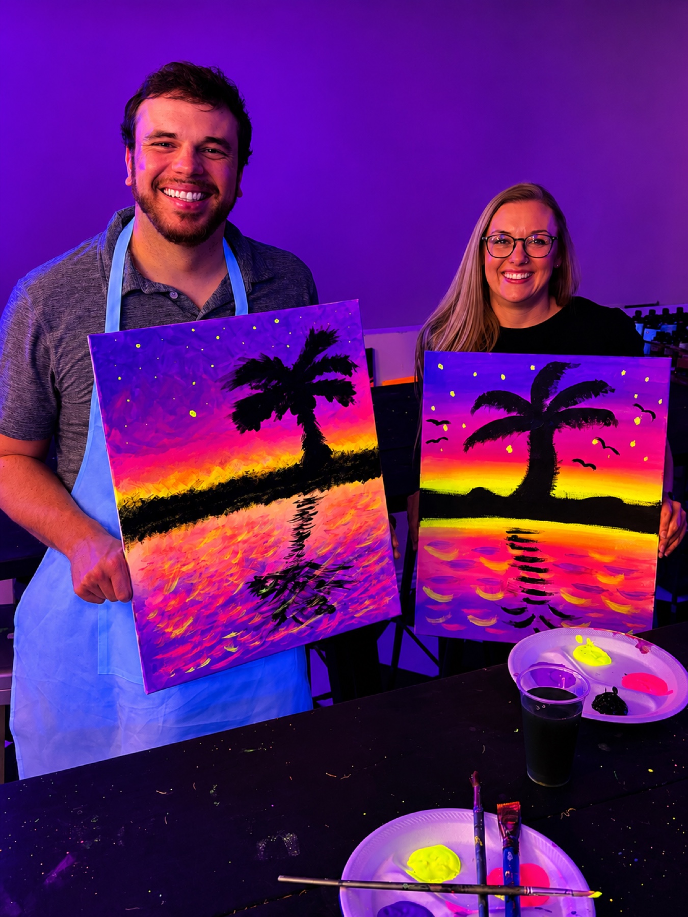 Couple holding bright tropical sunset canvases after a UV painting class at Studio 786 Houston