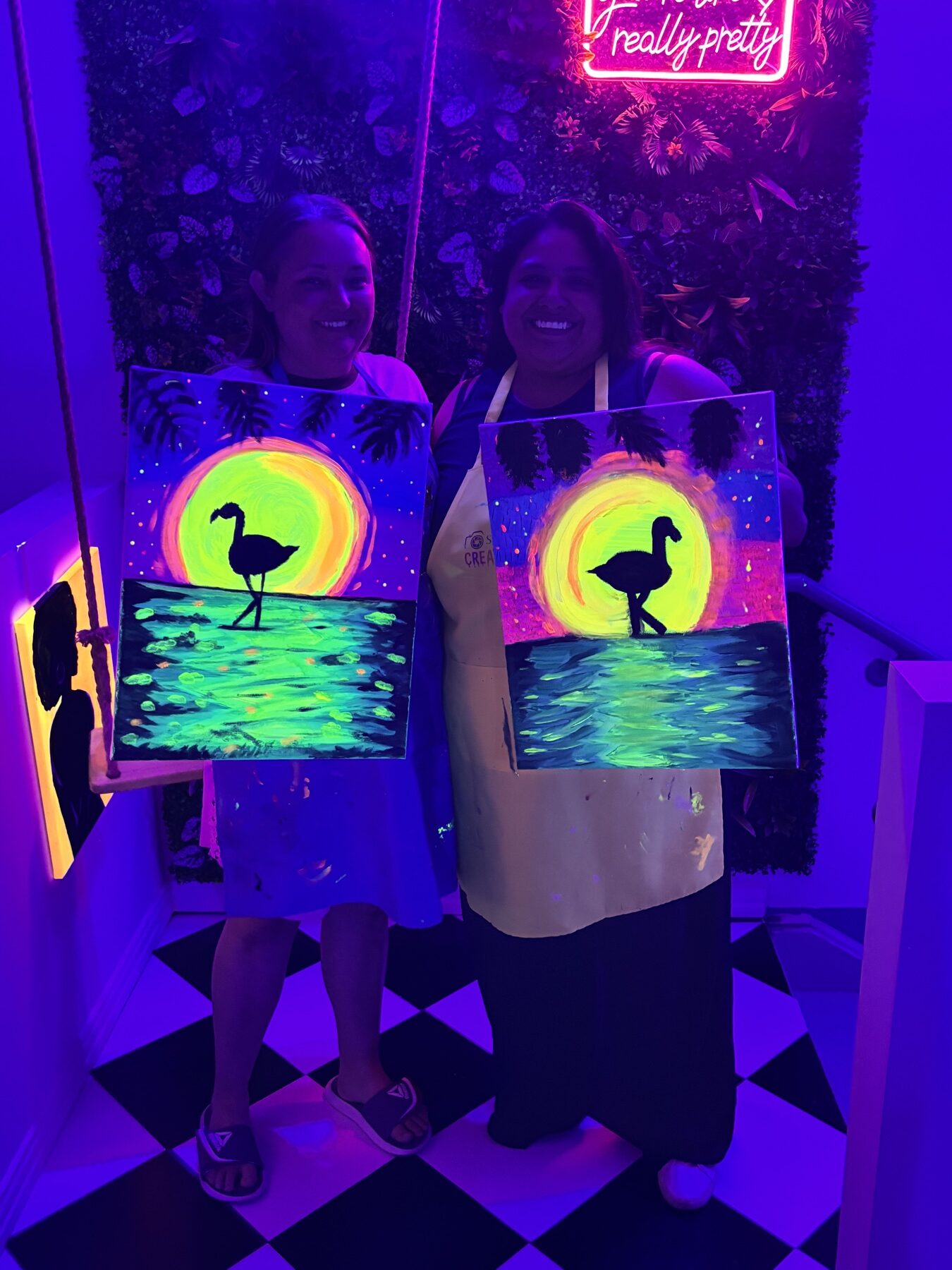 Friends holding bright flamingo canvases after a UV glow painting class in Houston