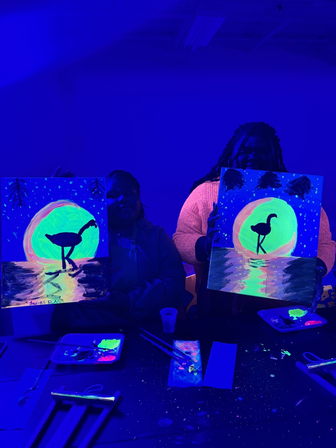 Guests holding finished neon flamingo paintings under UV lights at Studio 786 Houston